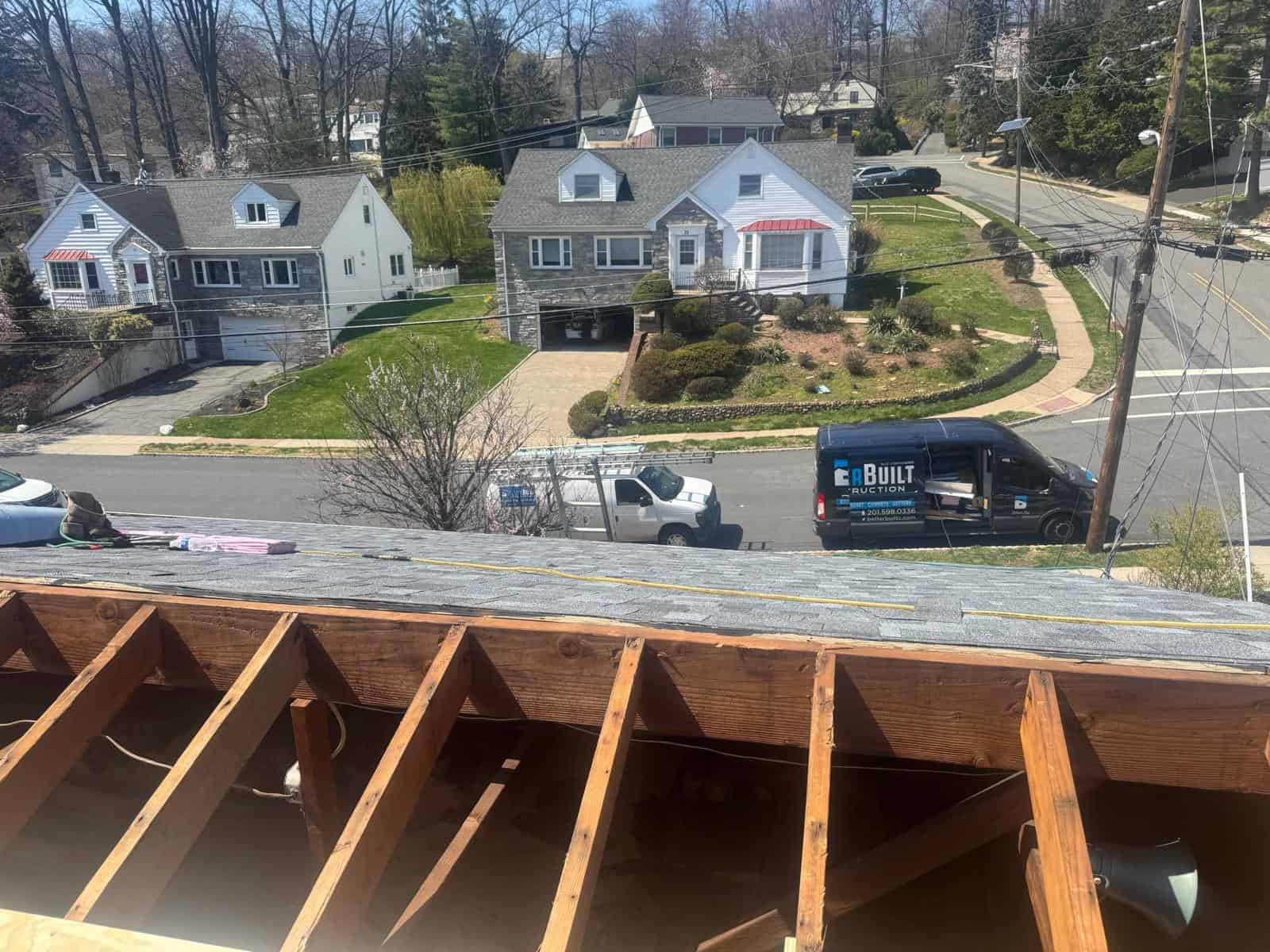 Roof rafters exposed at the edge of a New Jersey roof during repair work, with Better Build Construction vans on the street below